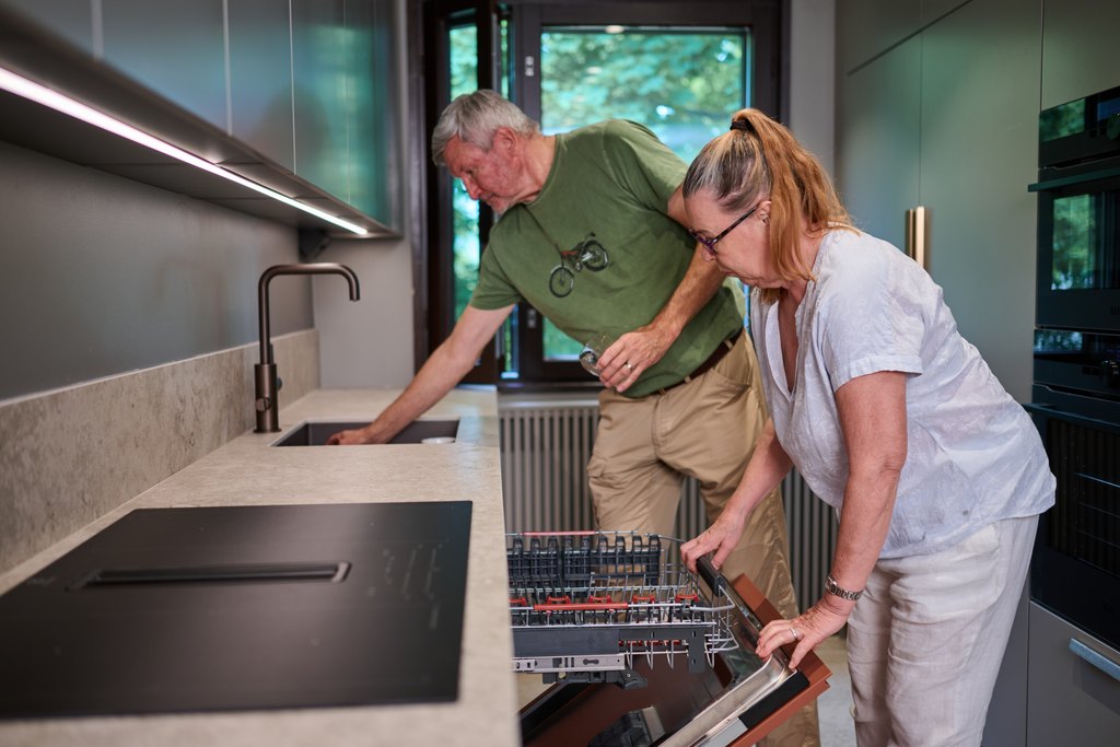 Kaksi iäkästä henkilöä modernissa keittiössä. Toinen kumartuu tarkastelemaan avattua astianpesukonetta ja toinen katsoo vieressä tiskialtaan luona. Keittiö on valoisa ja esteetön.