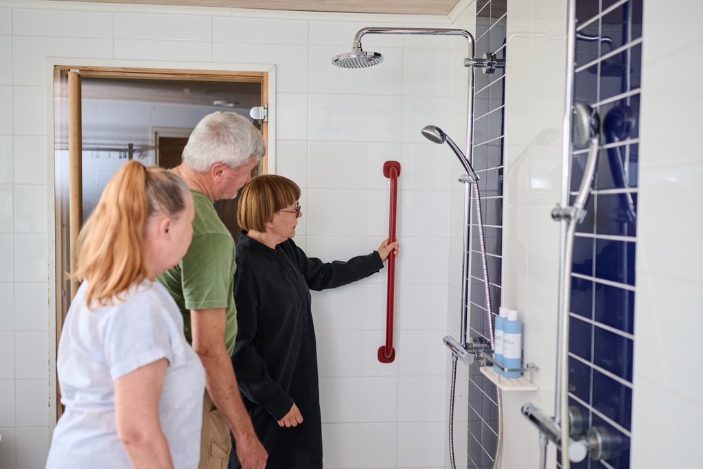 Kolme iäkästä henkilöä kylpyhuoneessa. Yksi heistä esittelee seinään asennettavaa tukikaidetta, kun kaksi muuta seuraa ja kuuntelee. Kylpyhuone on vaalea ja tarkoitettu esteettömäksi ja turvalliseksi.