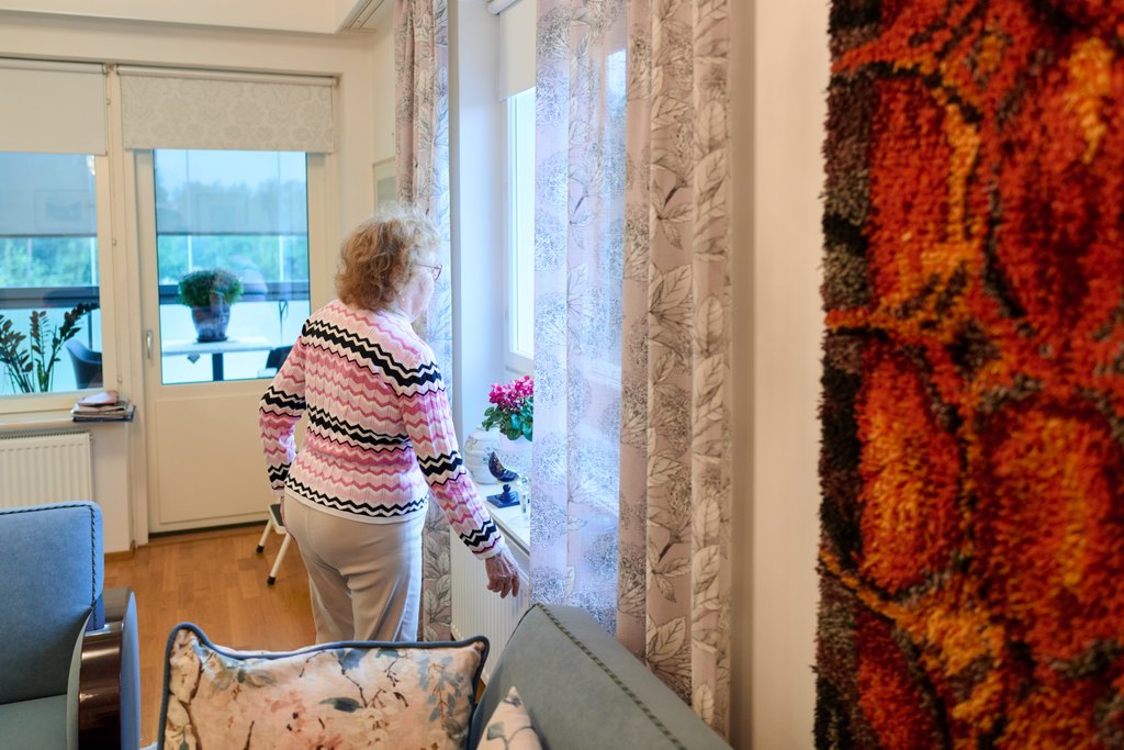 Iäkäs henkilö seisoo olohuoneessa ikkunan äärellä ja katsoo ulos. Huone on kodikas ja valoisa, ja sisustuksessa näkyy sohva, verhot sekä kasveja ikkunalaudalla.