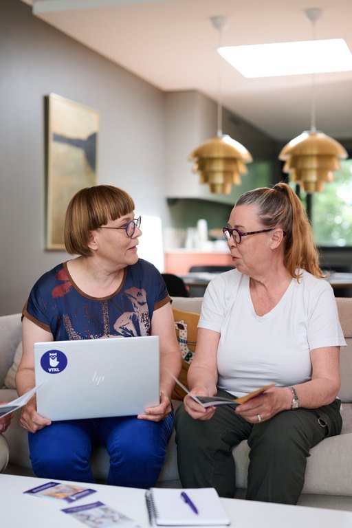 Kaksi naista istuu sohvalla ja keskustelee. Toisella on sylissään kannettava tietokone ja toisella esitteitä kädessään. Pöydällä on muistivihko ja kynä, ja taustalla näkyy kodin olohuonetila valaisimineen.
