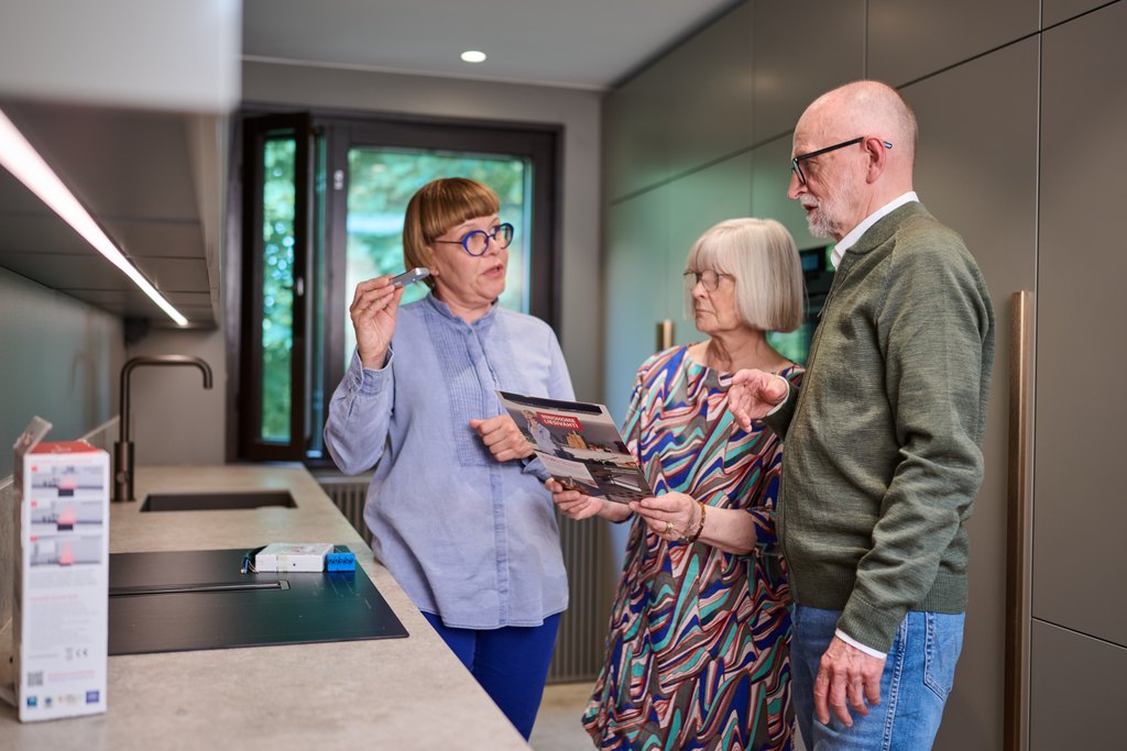 Kolme aikuista henkilöä keskustelee modernissa keittiössä. Yksi esittelee pientä laitetta kädessään, ja kaksi muuta katsoo esitettä. Keittiössä on vaaleat tasot ja taustalla ikkuna, josta tulee luonnonvaloa.