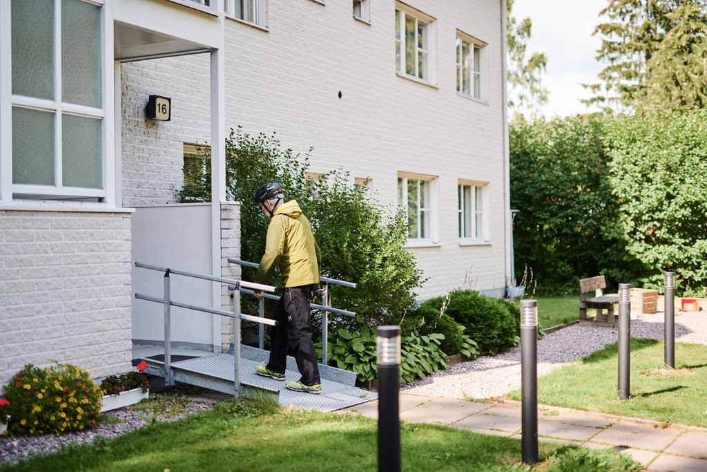 Henkilö nousee loivaa luiskaa pitkin kerrostalon sisäänkäynnille. Sisäänkäynnillä on kaiteet ja ympärillä piha-alue istutuksineen ja penkkeineen.