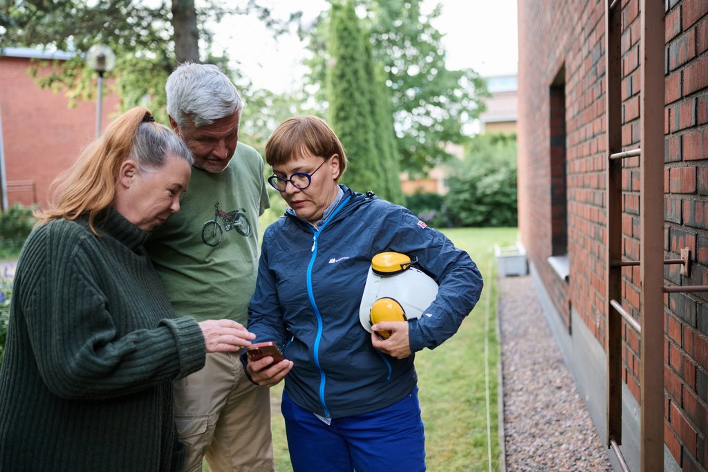 Kolme aikuista seisoo ulkona kerrostalon pihalla ja katsoo yhdessä puhelinta. Yhdellä heistä on kädessään mittauslaite ja kypärä, ja vieressä näkyy talon seinä sekä esteettömyyteen liittyviä rakenteita. Tilanne viittaa asumisen kartoitukseen tai neuvontaan.