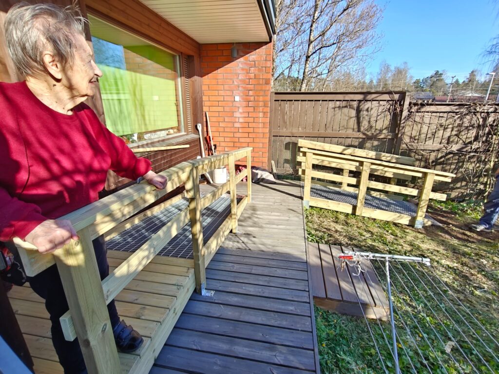 Iäkäs nainen seisoo terassilla uuden puisen luiskan vieressä ja pitää kiinni kaiteesta. Aurinkoisessa pihassa näkyy rakenteilla oleva esteetön kulkureitti sekä rakennustarvikkeita. Luiska helpottaa liikkumista kodin sisäänkäynnin ja pihan välillä.