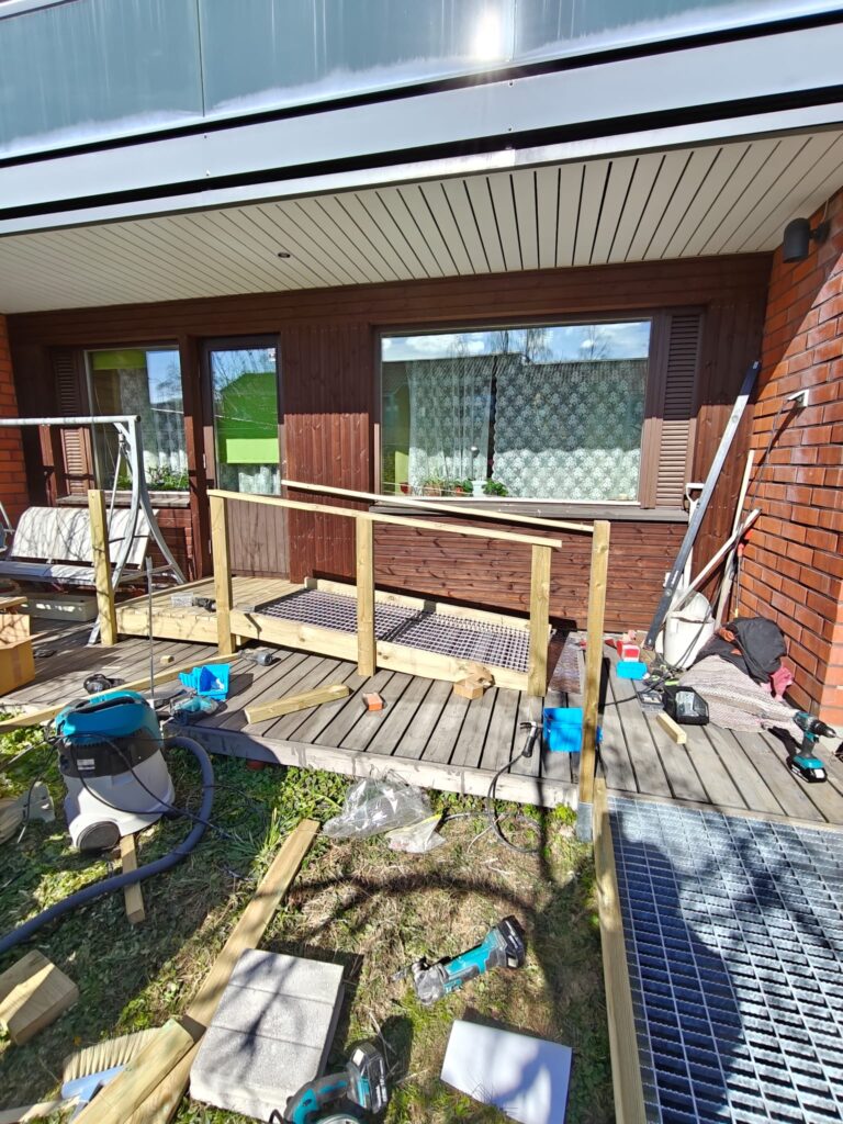 Terassin edustalla rakenteilla oleva puinen luiska tai kulkusilta omakotitalon sisäänkäynnille. Kuvassa näkyy rakennustarvikkeita, työkaluja ja keskeneräinen kaiderakenne aurinkoisessa pihassa. Luiska yhdistää pihan ja terassin helpottaen kulkua sisäänkäynnille.