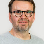 Lähikuva silmälasipäisestä henkilöstä vaaleanharmaassa t-paidassa. Hän katsoo kameraan ja hymyilee kevyesti. Taustana on vaalea, yksivärinen seinä.