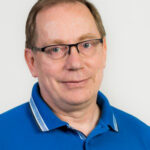 Lähikuva silmälasipäisestä henkilöstä, jolla on lyhyet vaaleanruskeat hiukset. Hän katsoo kameraan ja hymyilee hillitysti. Yllään hänellä on sininen kauluspaita. Taustana on vaalea, yksivärinen seinä.